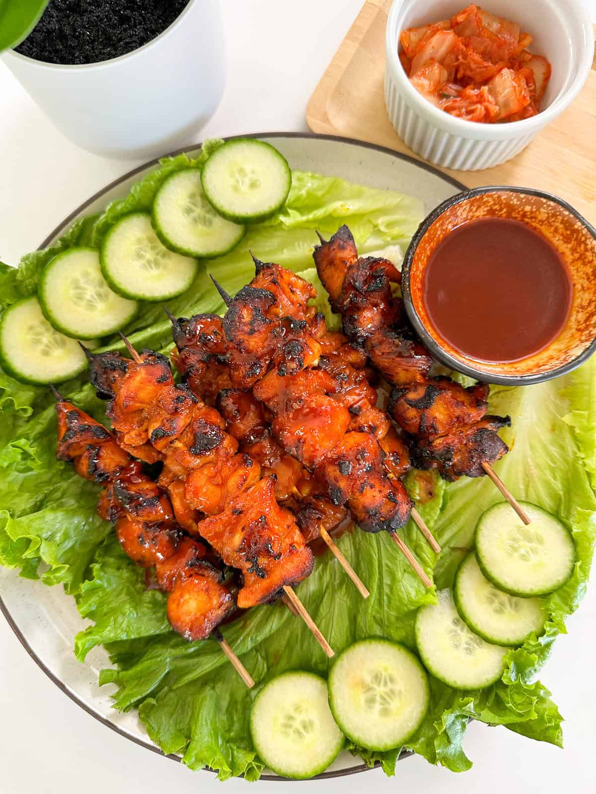 Spicy Gochujang Chicken Skewers coated in a vibrant red sauce are served on lettuce leaves with cucumber slices, a small bowl of dipping sauce, and a side of kimchi on a wooden board.