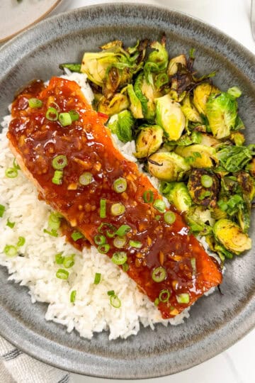 Sweet chili salmon on a bed of white rice with green onions on top and brussels sprouts to the side. The food is sitting in a dark colored plate. This is a top down view of the dish.