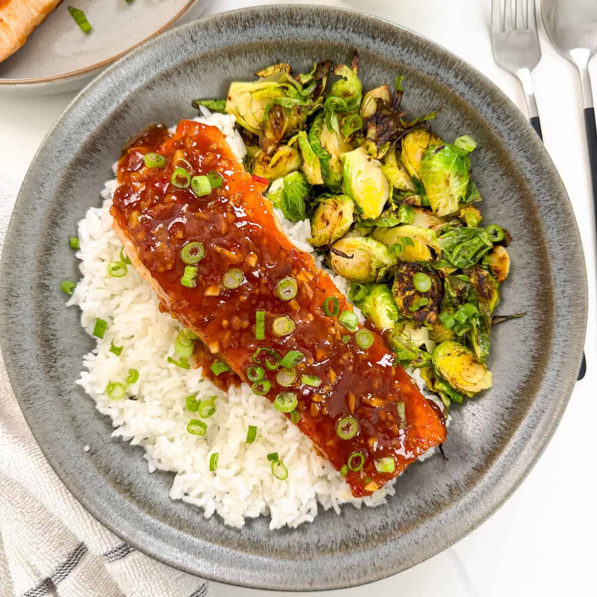 Sweet chili salmon on a bed of white rice with green onions on top and brussels sprouts to the side. The food is sitting in a dark colored plate. This is a top down view of the dish.