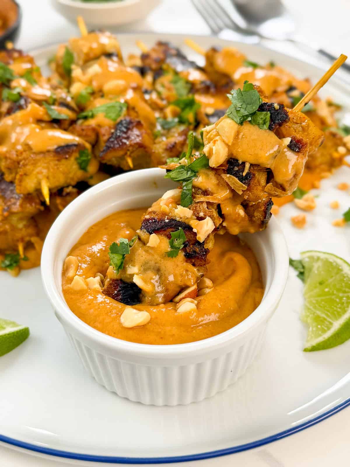 A Thai Chicken Satay with Peanut Sauce skewer, garnished with herbs and chopped peanuts, is dipped in a creamy orange sauce. More skewers, sauce, lime wedges, and herbs fill a white plate in the background.