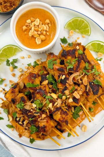 A plate of Thai Chicken Satay with Peanut Sauce features grilled chicken skewers topped with chopped peanuts and cilantro, served with lime wedges and a small bowl of peanut sauce. A spoon and napkin are beside the plate.