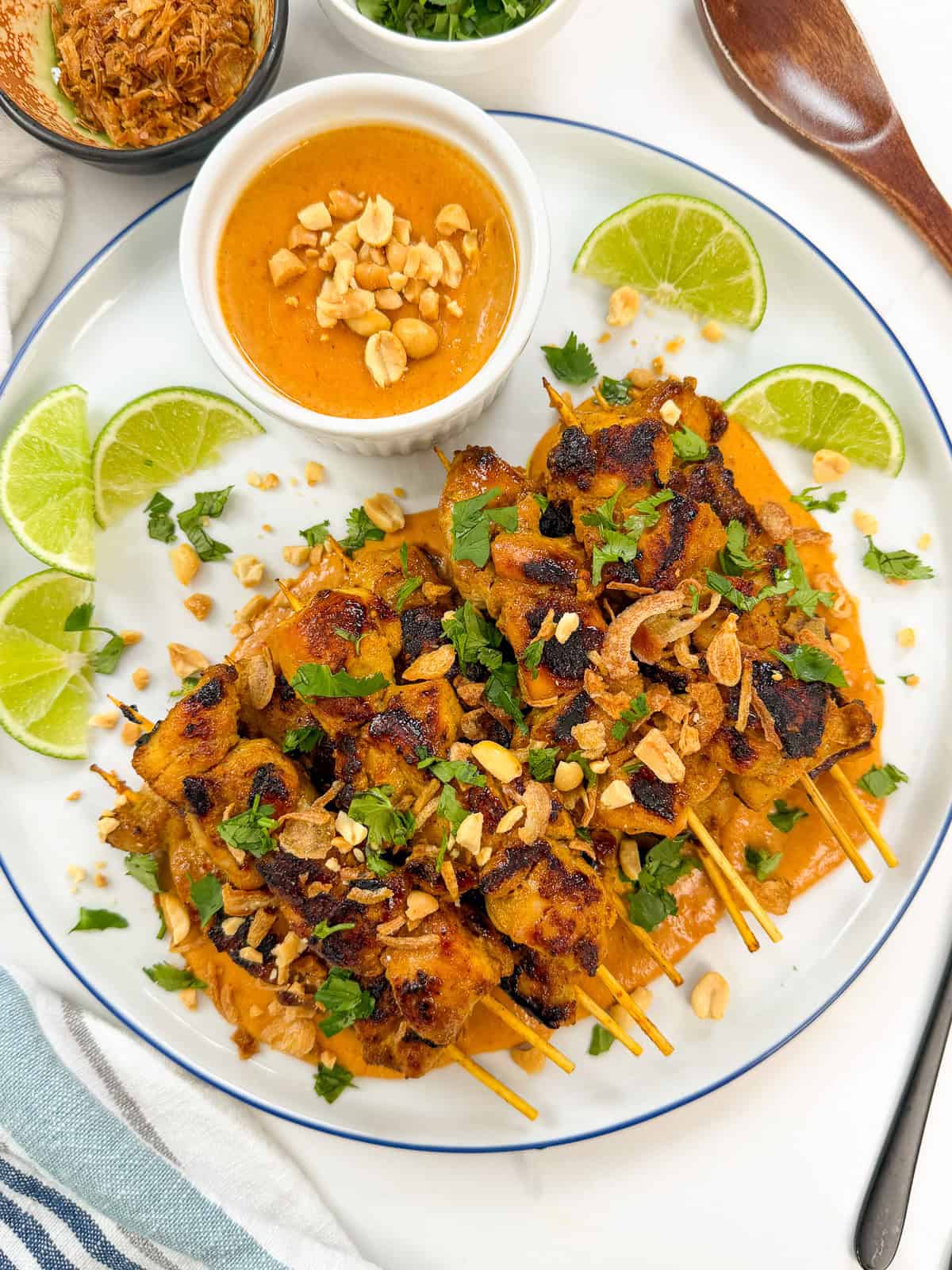 Thai Chicken Satay with Peanut Sauce: Grilled chicken skewers topped with chopped peanuts, herbs, and fried shallots on a white plate, served with creamy peanut sauce and lime wedges. A wooden spoon and small bowls of garnishes are nearby.
