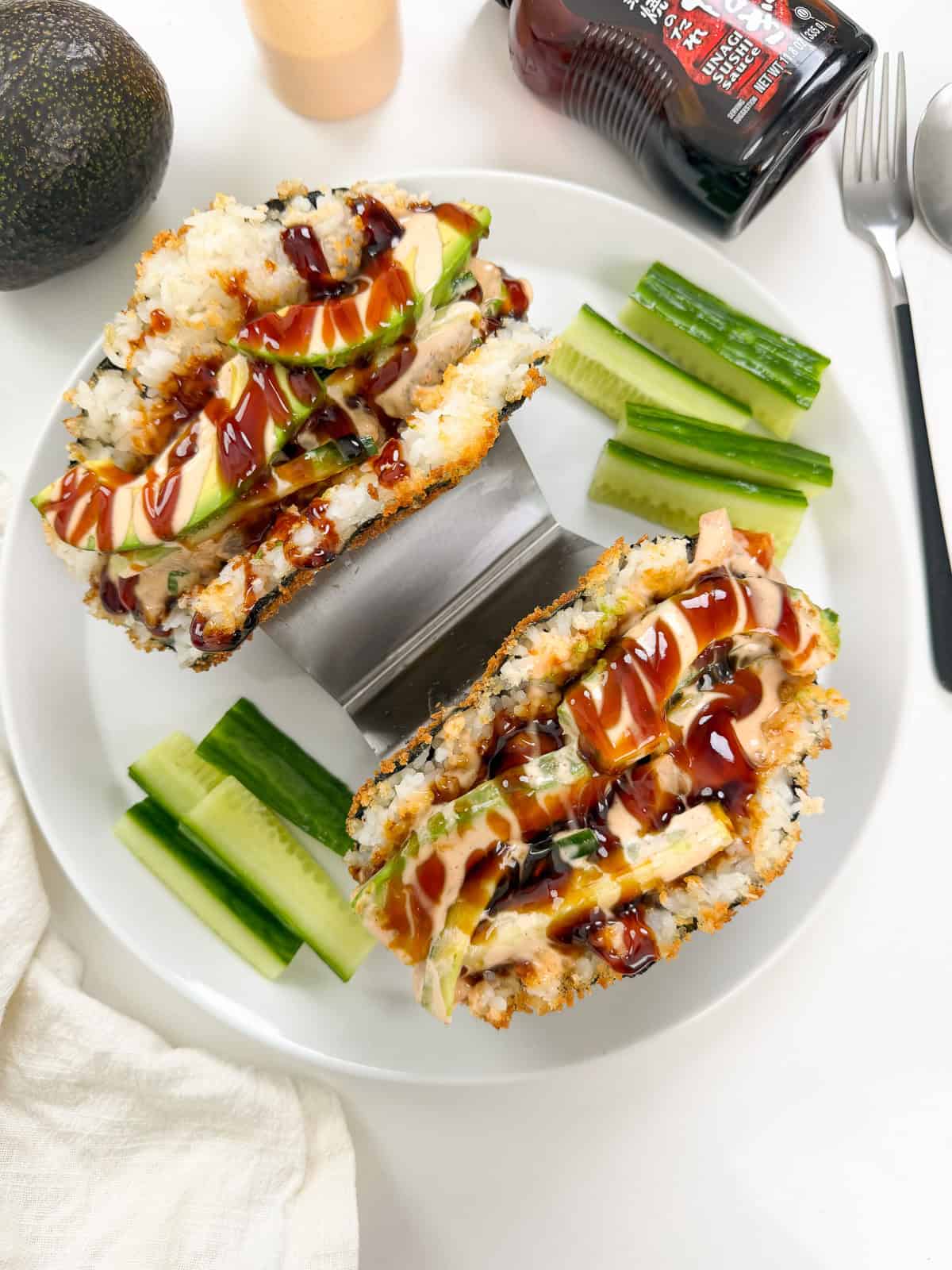 Two sushi sandwiches, inspired by Viral Sushi Tacos, are filled with rice, avocado, cucumber, and sauce on a plate, garnished with sliced cucumber and a bottle of soy sauce nearby. A fork and a halved avocado sit at the side.
