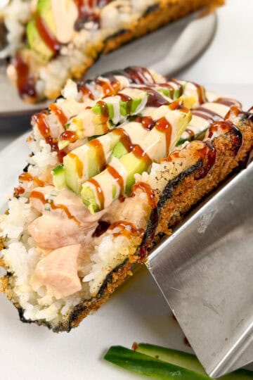 A viral sushi taco with a crispy seaweed shell, filled with rice, raw fish, sliced avocado, drizzled in creamy and dark sauces, served on a metal taco holder with cucumber slices nearby.