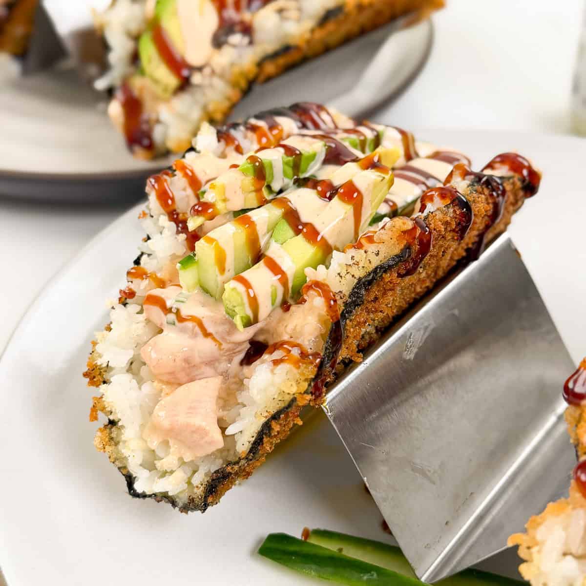 A viral sushi taco with a crispy seaweed shell, filled with rice, raw fish, sliced avocado, drizzled in creamy and dark sauces, served on a metal taco holder with cucumber slices nearby.