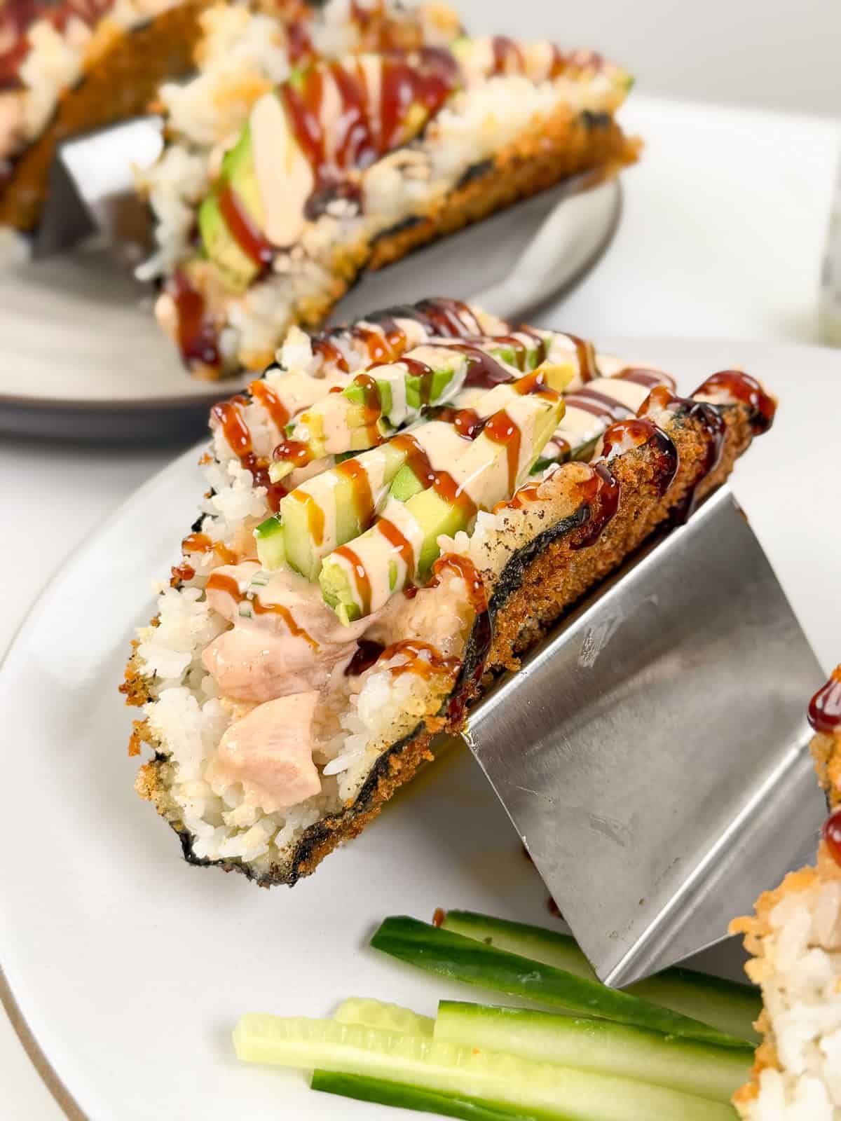 A crispy Viral Sushi Tacos shell filled with rice, raw fish, sliced avocado, and cucumber, drizzled with sauces and served on a metal holder with cucumber sticks on the side.