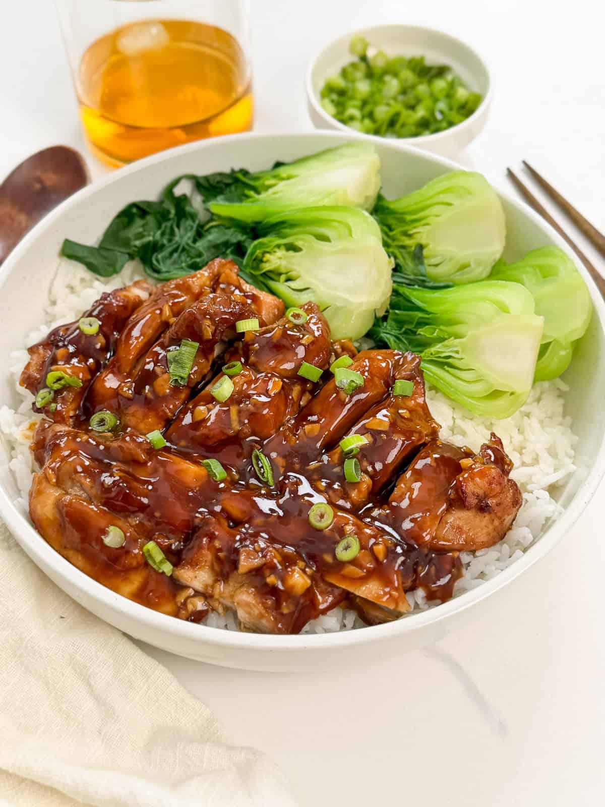 A bowl of white rice topped with glazed bourbon chicken, chopped green onions, steamed bok choy, and leafy greens, with a glass of amber beverage and a small bowl of sliced green onions in the background.