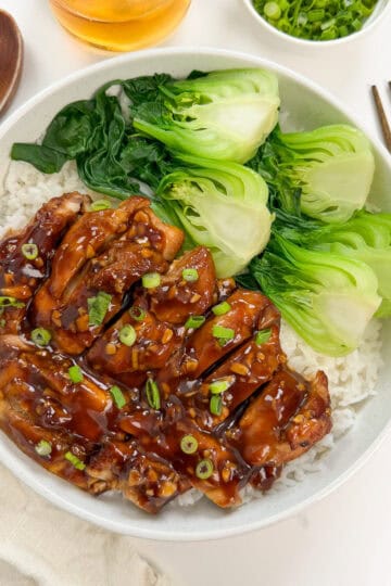 A bowl of rice topped with tender Bourbon Chicken slices, glazed to perfection, steamed bok choy, and leafy greens, garnished with chopped green onions. Chopsticks and a wooden spoon are placed beside the bowl.
