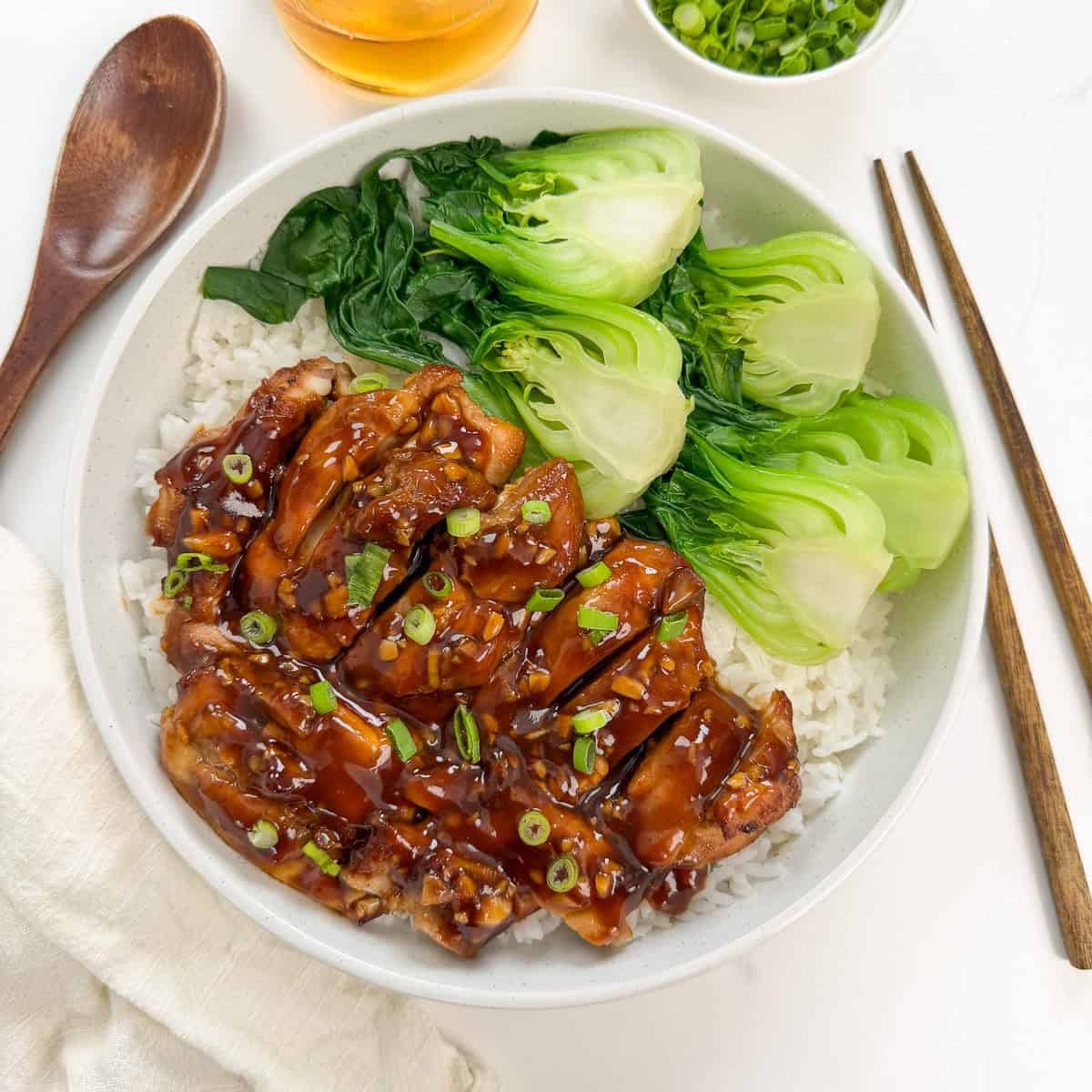 A bowl of rice topped with tender Bourbon Chicken slices, glazed to perfection, steamed bok choy, and leafy greens, garnished with chopped green onions. Chopsticks and a wooden spoon are placed beside the bowl.