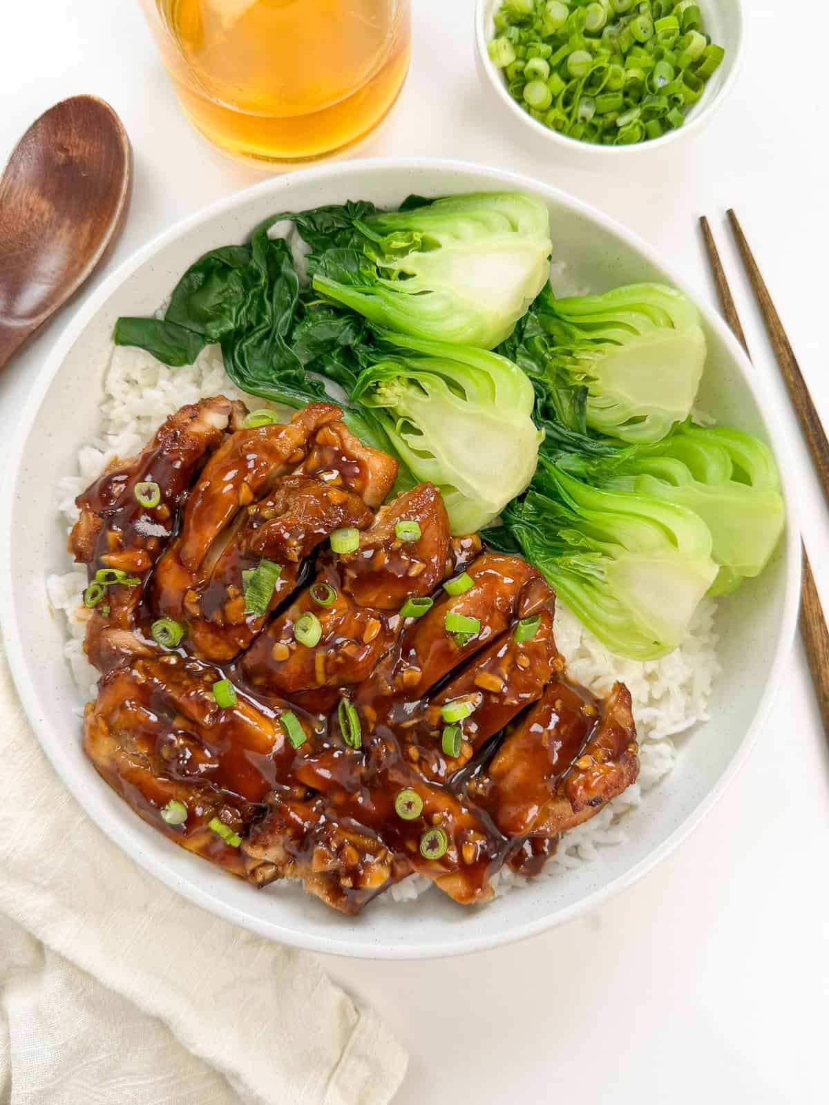 A bowl of rice topped with glazed bourbon chicken, sliced bok choy, steamed greens, and garnished with chopped green onions. Chopsticks, a wooden spoon, a napkin, and a bowl of green onions are nearby.