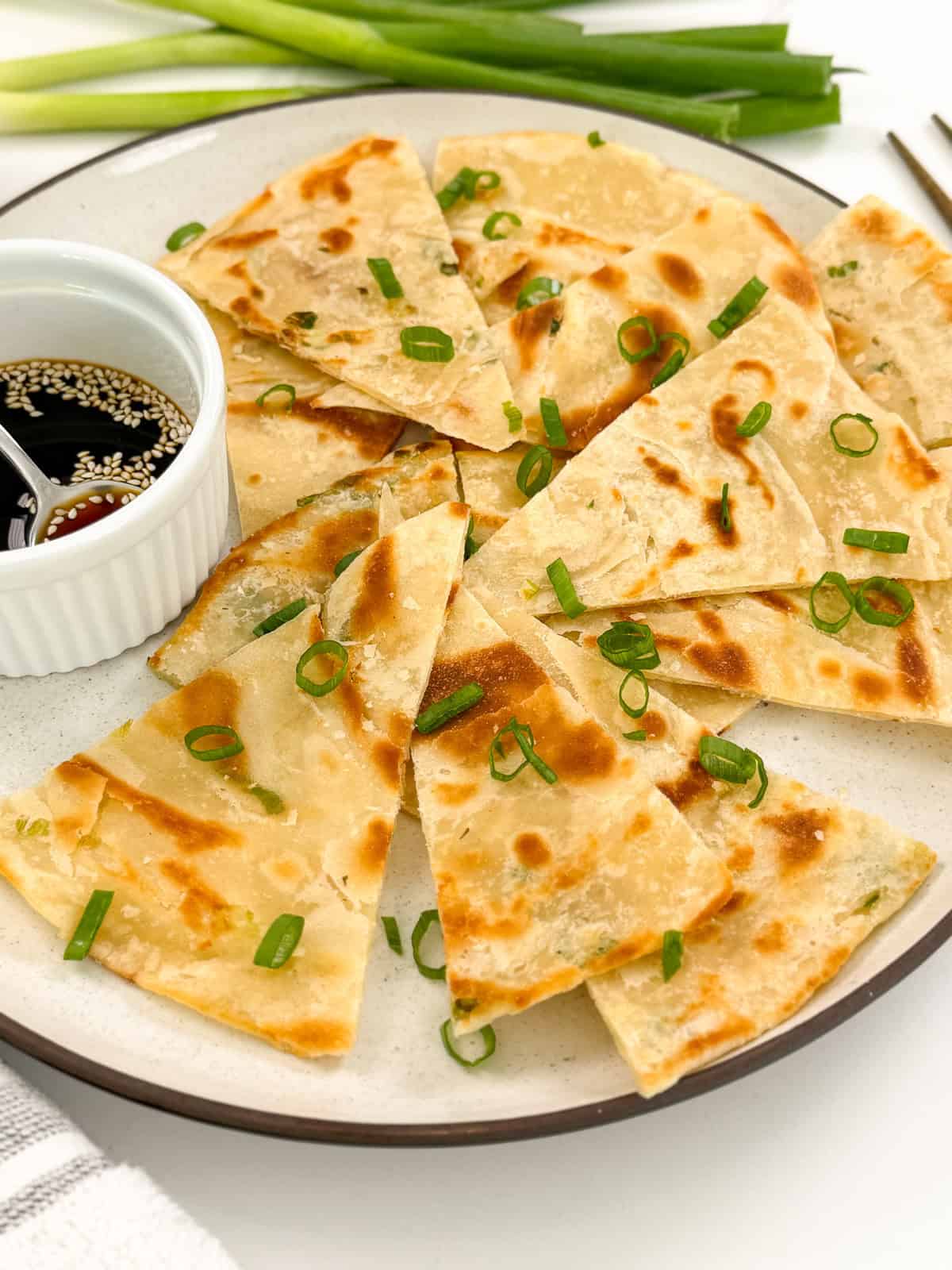 Chinese Scallion Pancakes are sliced and arranged on a plate, garnished with chopped green onions, and served with a small bowl of soy sauce topped with sesame seeds. Fresh green onions feature in the background.