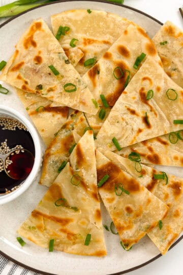 A plate of sliced Chinese Scallion Pancakes garnished with chopped green onions, served with a small bowl of soy dipping sauce topped with sesame seeds and a spoon. Chopsticks are placed beside the plate.