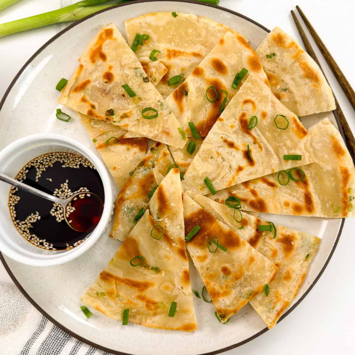A plate of sliced Chinese Scallion Pancakes garnished with chopped green onions, served with a small bowl of soy dipping sauce topped with sesame seeds and a spoon. Chopsticks are placed beside the plate.