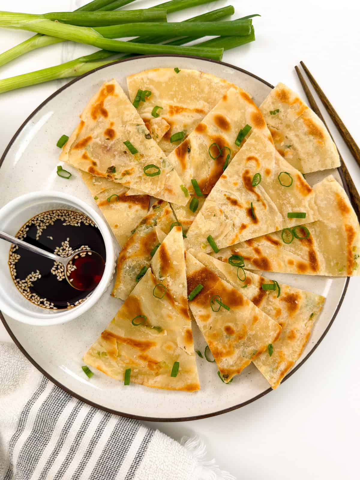 A plate of sliced Chinese Scallion Pancakes garnished with chopped green onions, served with a small bowl of soy dipping sauce with sesame seeds; green onions and chopsticks are nearby.