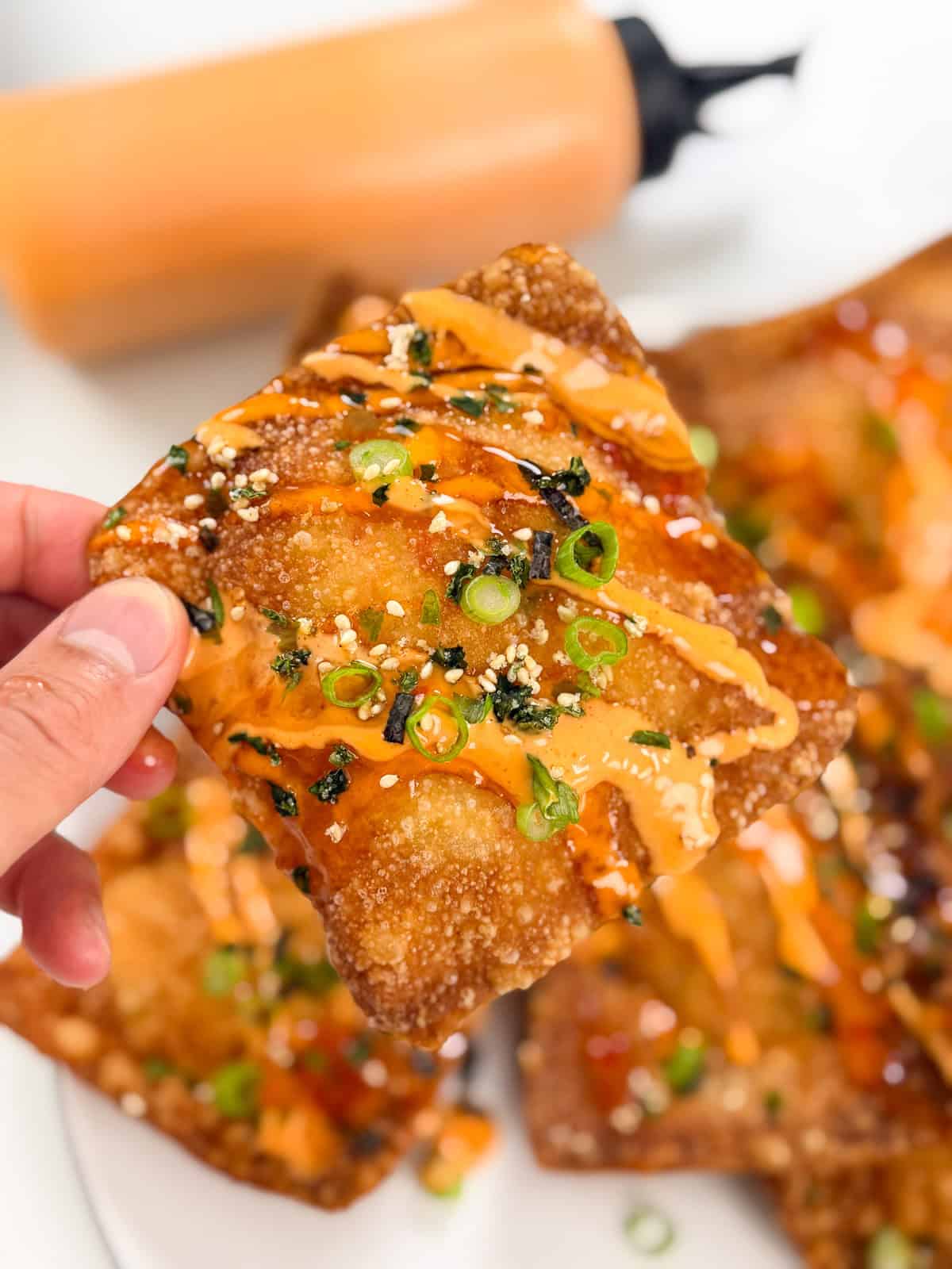 A hand holds a crispy, golden wonton topped with orange sauce, sliced green onions, sesame seeds, and seaweed flakes—these Crab Rangoon Nachos shine. More wontons and a squeeze bottle of sauce are visible in the background.