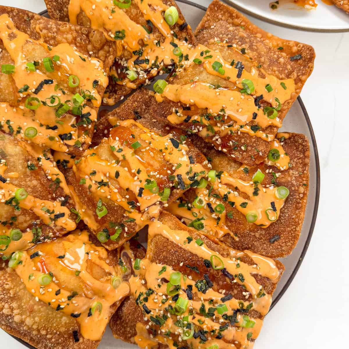 A plate of crispy, square wontons transforms into Crab Rangoon Nachos, topped with orange spicy mayo sauce, sesame seeds, chopped green onions, and bits of dried seaweed.