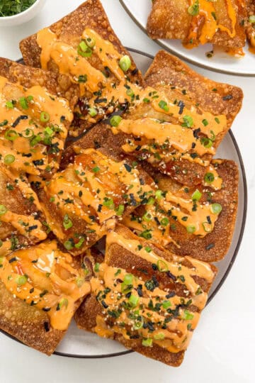 A plate of crispy, golden-brown wontons inspired by Crab Rangoon Nachos, garnished with chopped green onions, sesame seeds, and a drizzle of orange spicy mayo sauce. Another smaller plate with more wontons appears in the background.