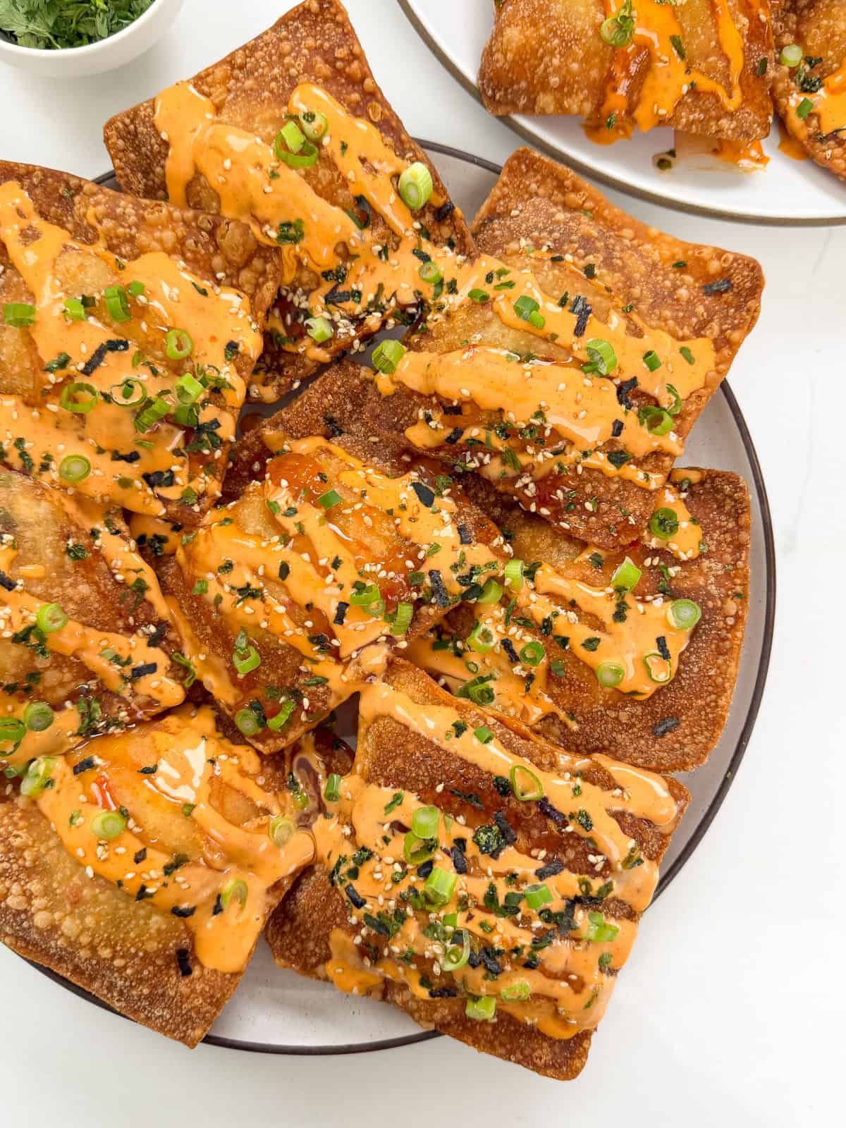 A plate of crispy, golden-brown wontons inspired by Crab Rangoon Nachos, garnished with chopped green onions, sesame seeds, and a drizzle of orange spicy mayo sauce. Another smaller plate with more wontons appears in the background.