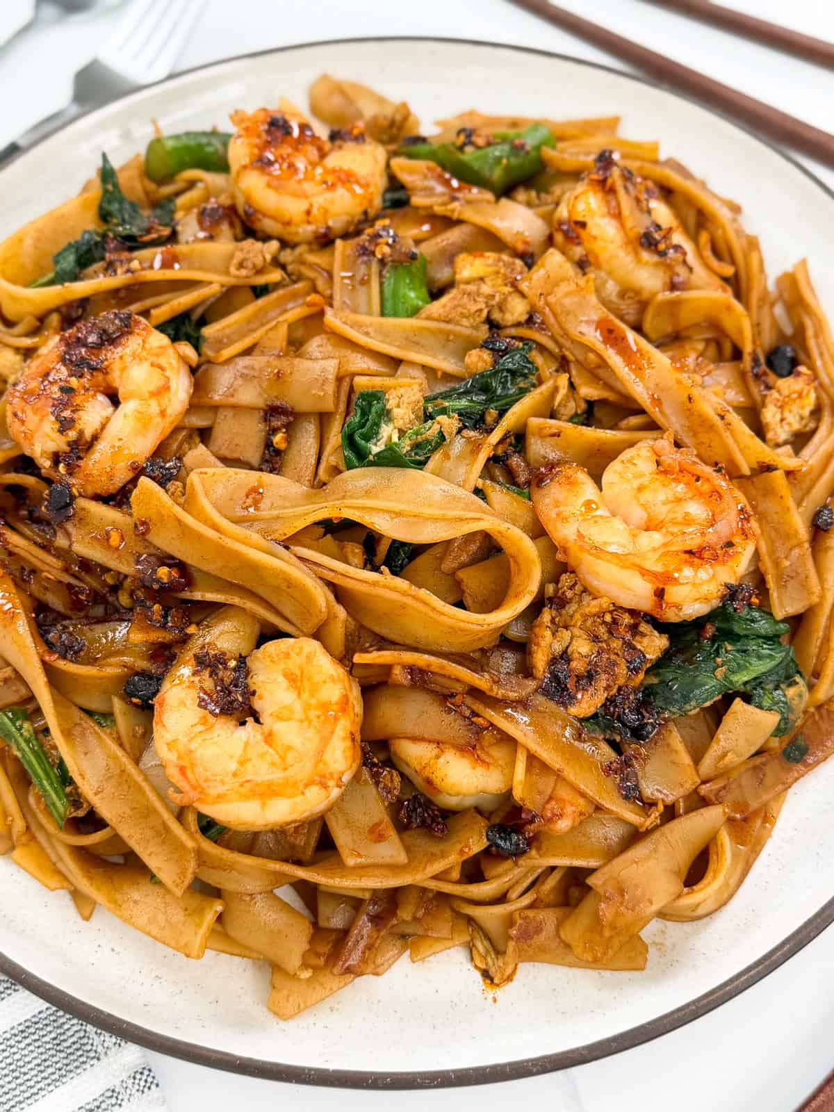 A plate of Easy Thai Pad See Ew features wide rice noodles stir-fried with shrimp, greens, and savory sauce, garnished with chili flakes. Chopsticks and a fork are placed nearby on a white tablecloth.