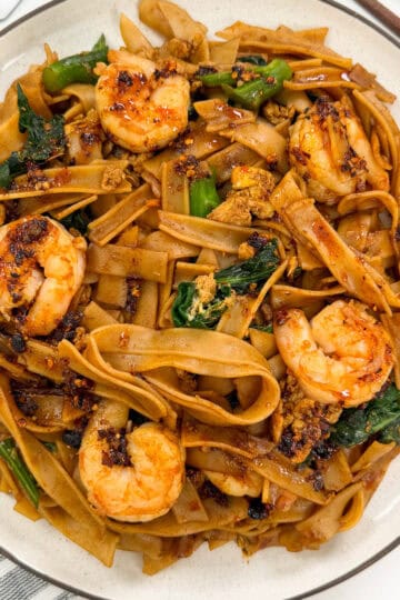 A plate of Easy Thai Pad See Ew features wide rice noodles stir-fried with shrimp, leafy greens, and savory sauce, garnished with chili flakes and scrambled egg bits. Chopsticks and a white napkin are nearby.