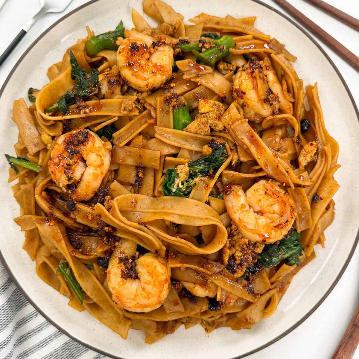 A plate of Easy Thai Pad See Ew features wide rice noodles stir-fried with shrimp, leafy greens, and savory sauce, garnished with chili flakes and scrambled egg bits. Chopsticks and a white napkin are nearby.
