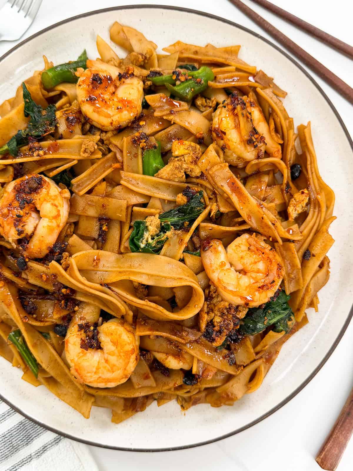 A plate of Easy Thai Pad See Ew features stir-fried wide rice noodles with shrimp, leafy greens, scrambled egg, and a savory brown sauce, garnished with chili flakes. Chopsticks are placed on the side.
