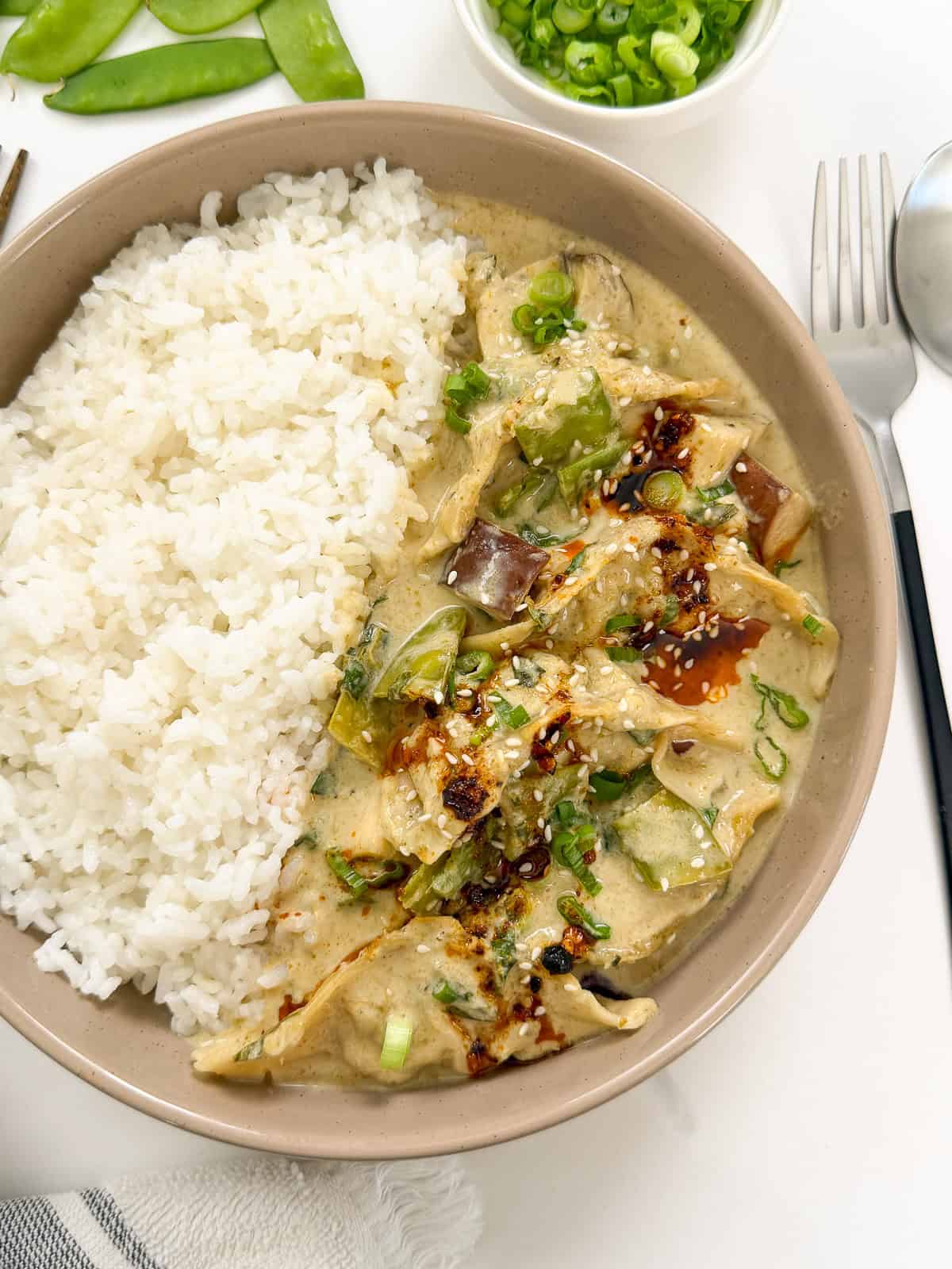 A beige bowl filled with white rice and creamy green curry with vegetables, inspired by a Green Curry Dumpling Bake, topped with sliced green onions and a drizzle of chili oil. A spoon, fork, and small bowl of green onions sit beside the dish.
