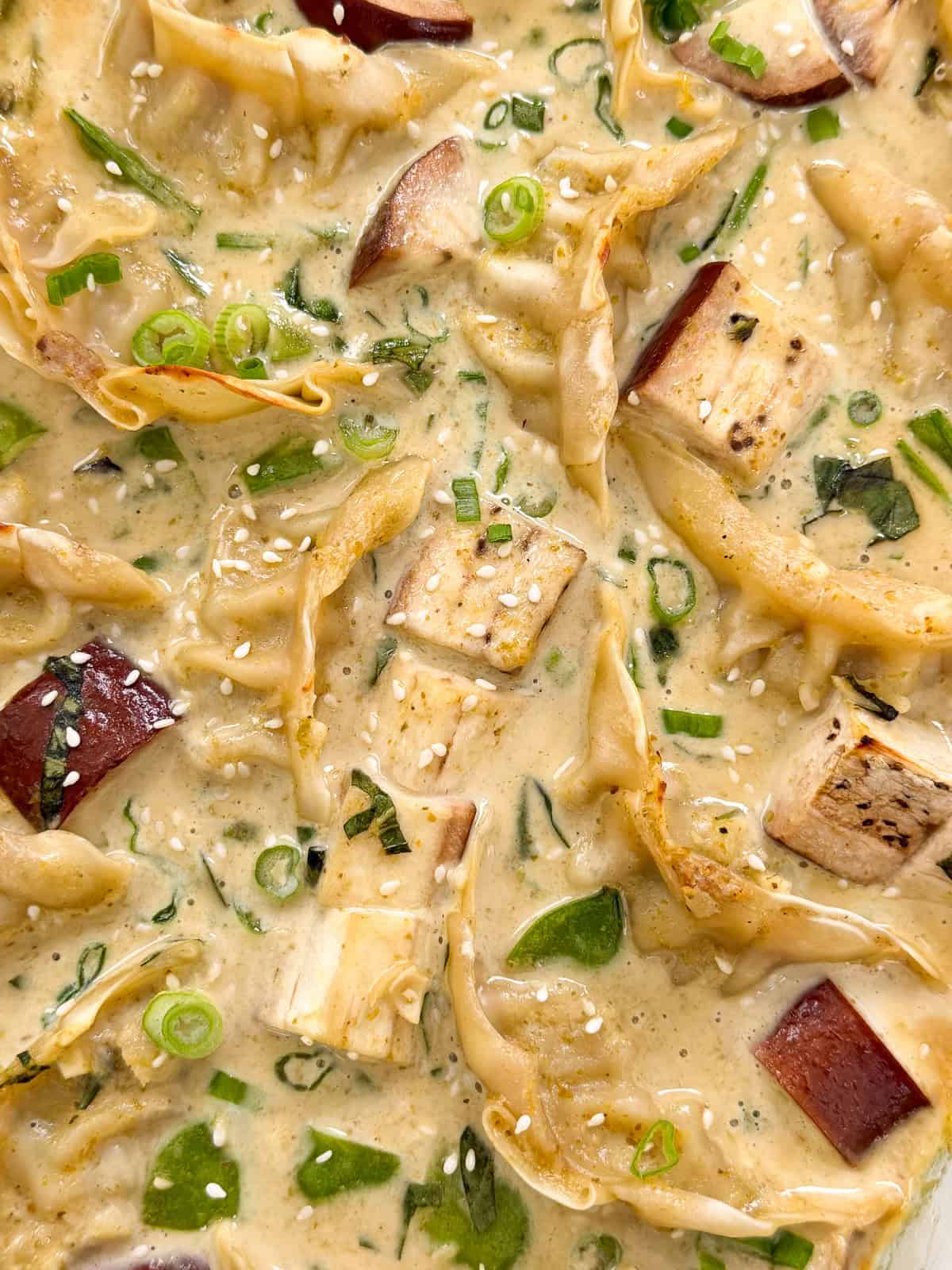 Close-up of creamy Green Curry Dumpling Bake with tofu cubes, chopped green onions, greens, and sesame seeds, all mixed in a rich, light-colored broth.