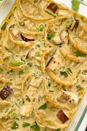 A glass baking dish filled with Green Curry Dumpling Bake in a creamy sauce, garnished with chopped green onions and chunks of eggplant, with a fork and napkin beside the dish.