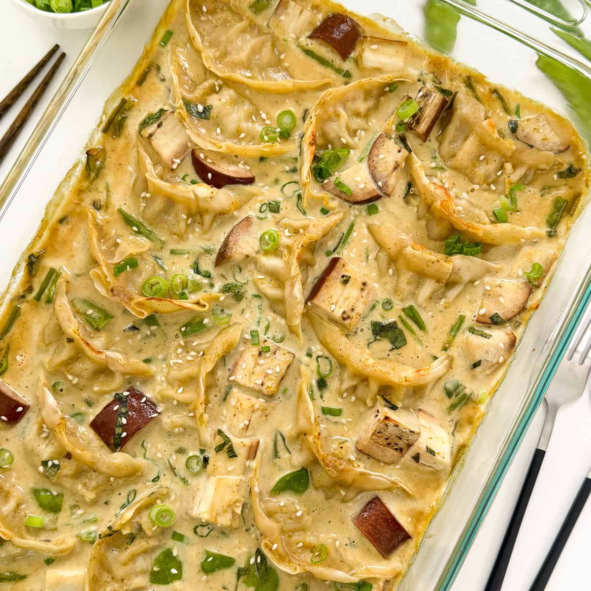 A glass baking dish filled with Green Curry Dumpling Bake in a creamy sauce, garnished with chopped green onions and chunks of eggplant, with a fork and napkin beside the dish.