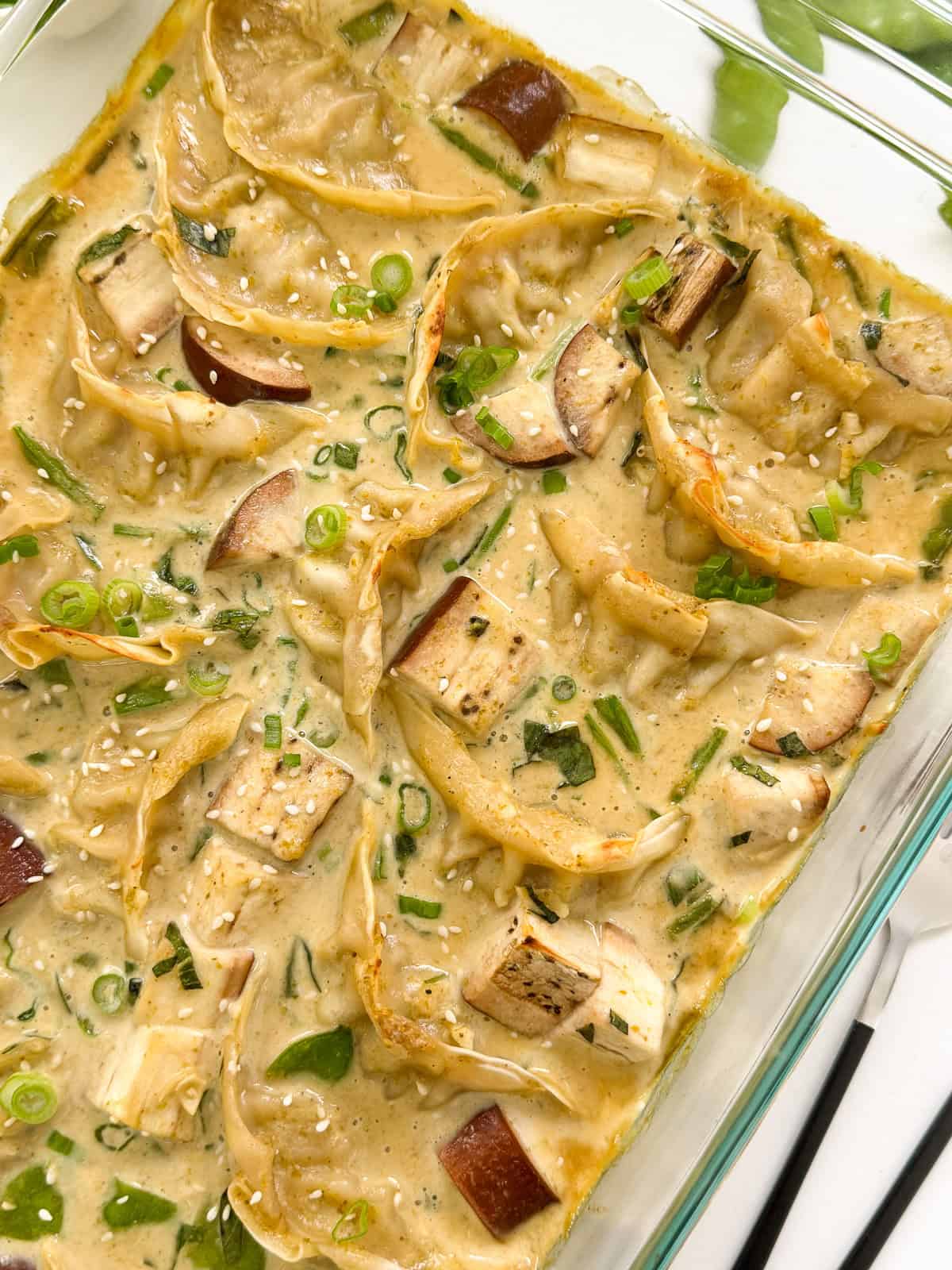 A glass baking dish filled with Green Curry Dumpling Bake in a creamy sauce, garnished with chopped green onions, sesame seeds, and cubes of eggplant and tofu.