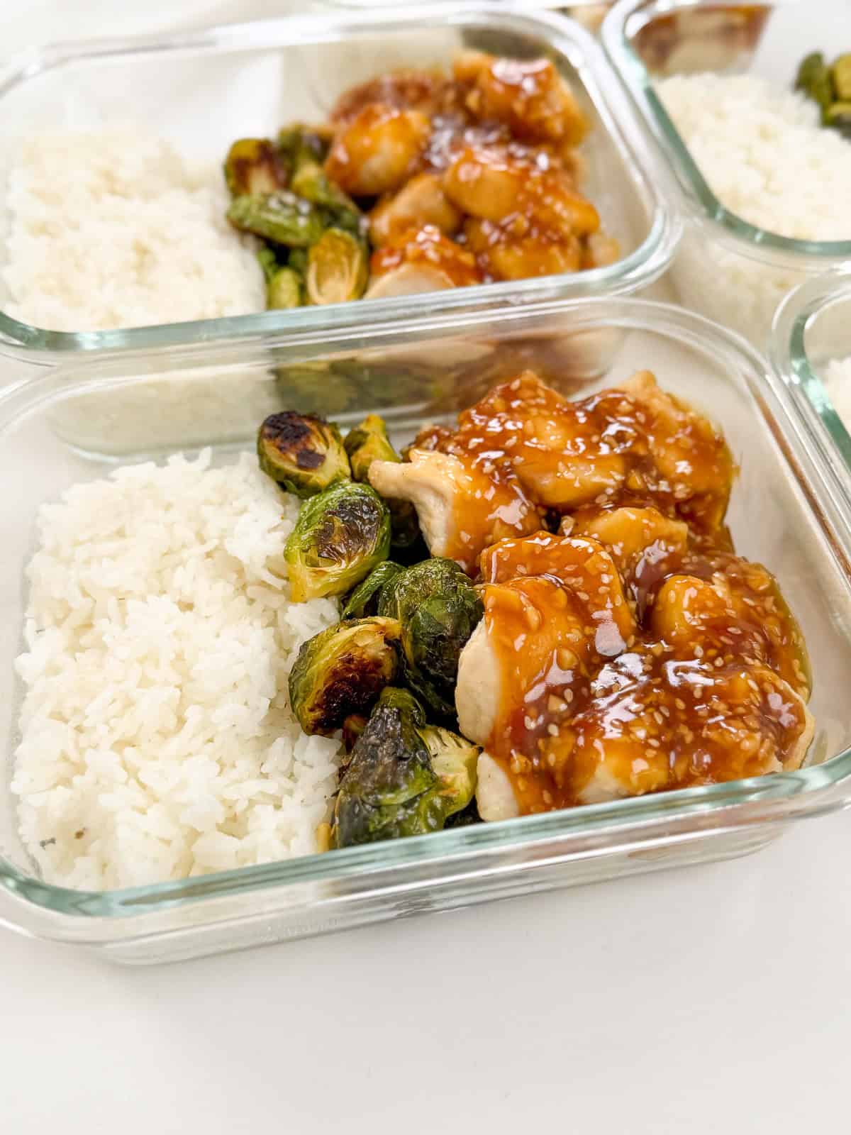 Glass meal prep containers filled with white rice, roasted Brussels sprouts, and pieces of chicken topped with a brown, sticky sauce. These Honey Sesame Chicken Meal Prep Bowls are arranged side by side on a white surface.
