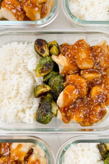 A glass meal prep container with white rice, roasted Brussels sprouts, and chicken pieces topped with a brown sesame sauce—perfect for Honey Sesame Chicken Meal Prep Bowls. Other similar containers are partially visible around it.