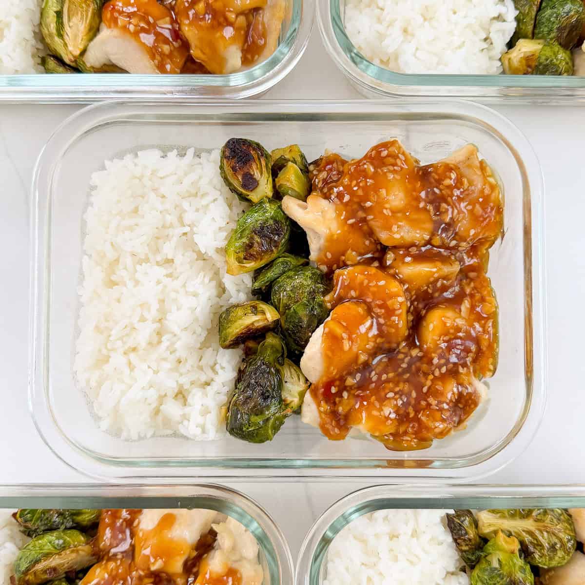 A glass meal prep container with white rice, roasted Brussels sprouts, and chicken pieces topped with a brown sesame sauce—perfect for Honey Sesame Chicken Meal Prep Bowls. Other similar containers are partially visible around it.