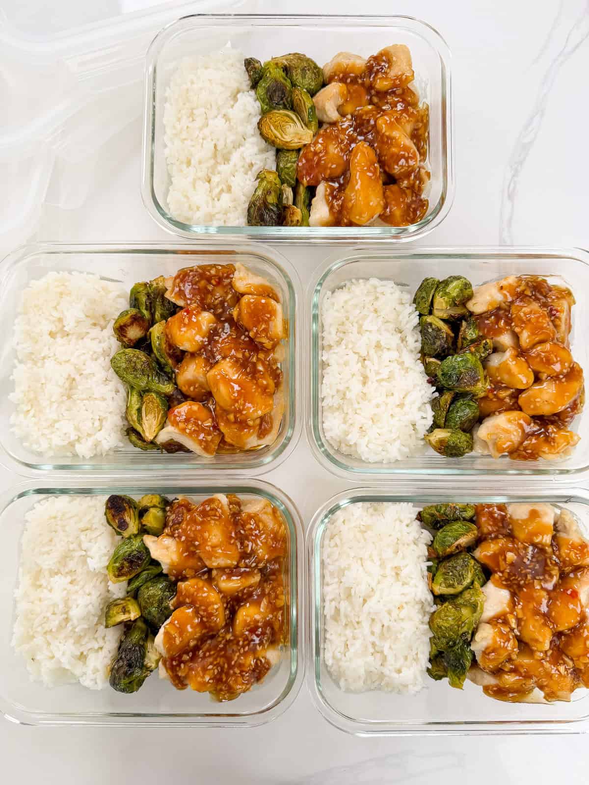Five glass meal prep containers filled with white rice, roasted Brussels sprouts, and Honey Sesame Chicken Meal Prep Bowls arranged side by side on a white surface.