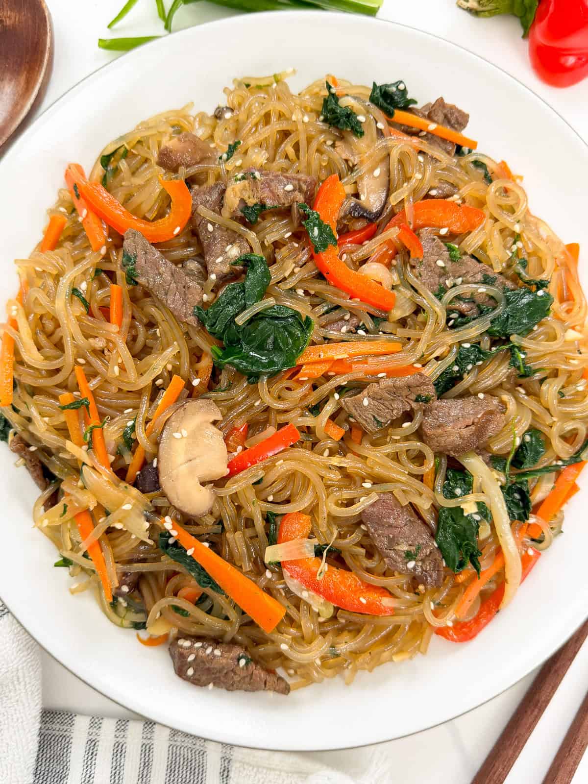A plate of Japchae (Korean Glass Noodle Stir-Fry) with beef, spinach, red bell peppers, mushrooms, carrots, and sesame seeds, all garnished and served on a white plate.