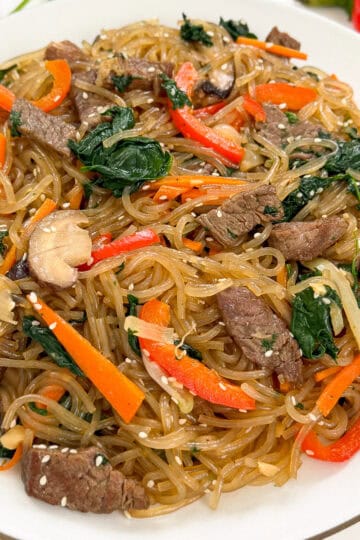 A plate of Japchae (Korean Glass Noodle Stir-Fry) with beef strips, carrots, spinach, mushrooms, onions, and sesame seeds, all perfectly stir-fried together.