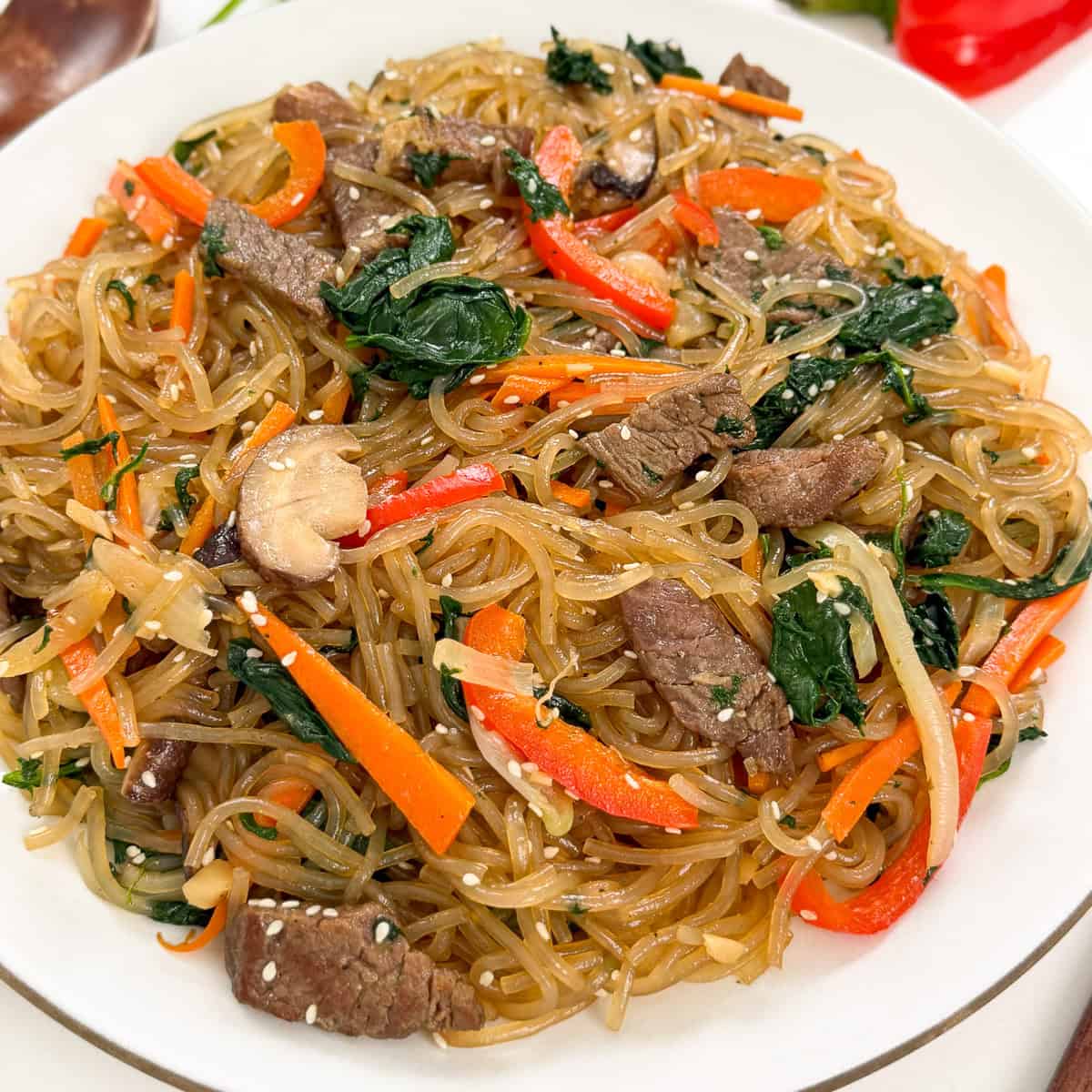 A plate of Japchae (Korean Glass Noodle Stir-Fry) with beef strips, carrots, spinach, mushrooms, onions, and sesame seeds, all perfectly stir-fried together.