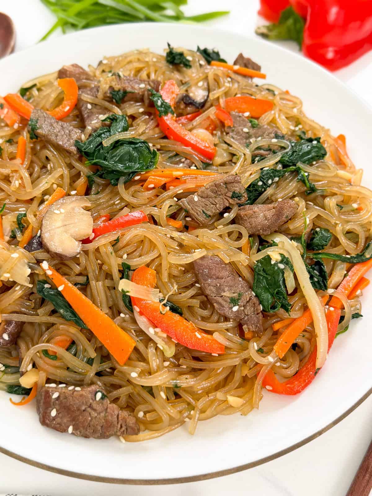 A plate of Japchae (Korean Glass Noodle Stir-Fry) with beef, spinach, carrots, red bell peppers, mushrooms, onions, and sesame seeds is beautifully served on a white dish.