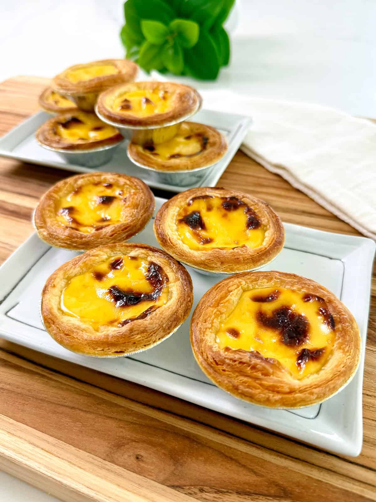 Six Portuguese Egg Tarts with golden, caramelized tops and flaky crusts are displayed on two rectangular plates atop a wooden board, with a green plant and white napkin in the background.