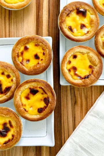 Golden Portuguese Egg Tarts with flaky crusts and creamy custard filling are arranged on two white rectangular plates atop a wooden table, with a light-colored napkin beside them.
