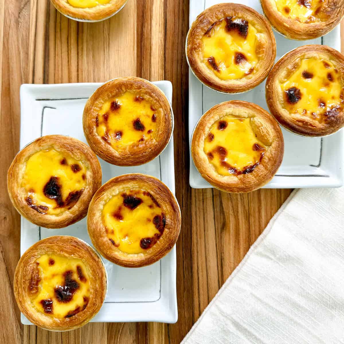 Golden Portuguese Egg Tarts with flaky crusts and creamy custard filling are arranged on two white rectangular plates atop a wooden table, with a light-colored napkin beside them.