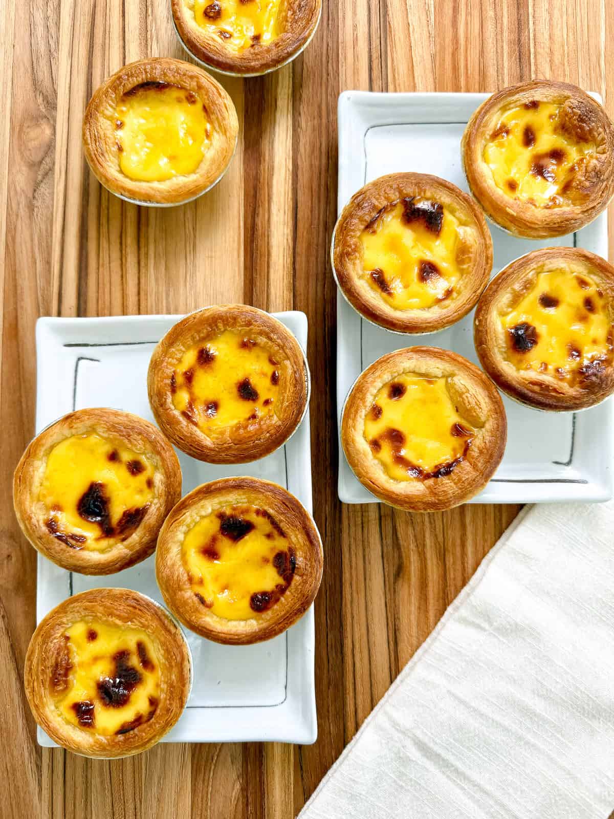 A top view of several golden Portuguese Egg Tarts with caramelized tops arranged on white rectangular plates, placed on a wooden surface next to a white cloth napkin.