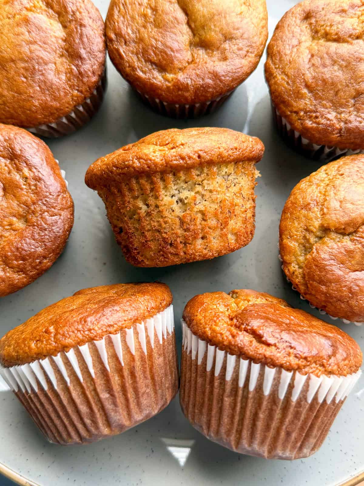 A close-up of several golden brown Protein Tahini Banana Muffins on a plate, with one muffin turned on its side to show its texture. Each muffin is wrapped in a white paper liner.