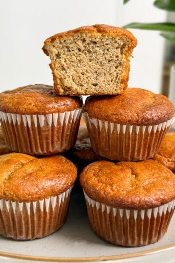 A plate of six Protein Tahini Banana Muffins in paper liners, with one muffin cut in half and placed on top to reveal its moist, textured inside. A green potted plant is blurred in the background.