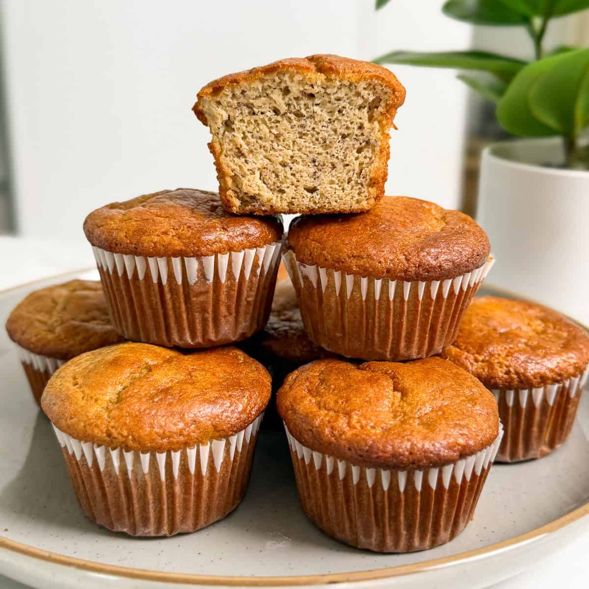 A plate of six Protein Tahini Banana Muffins in paper liners, with one muffin cut in half and placed on top to reveal its moist, textured inside. A green potted plant is blurred in the background.