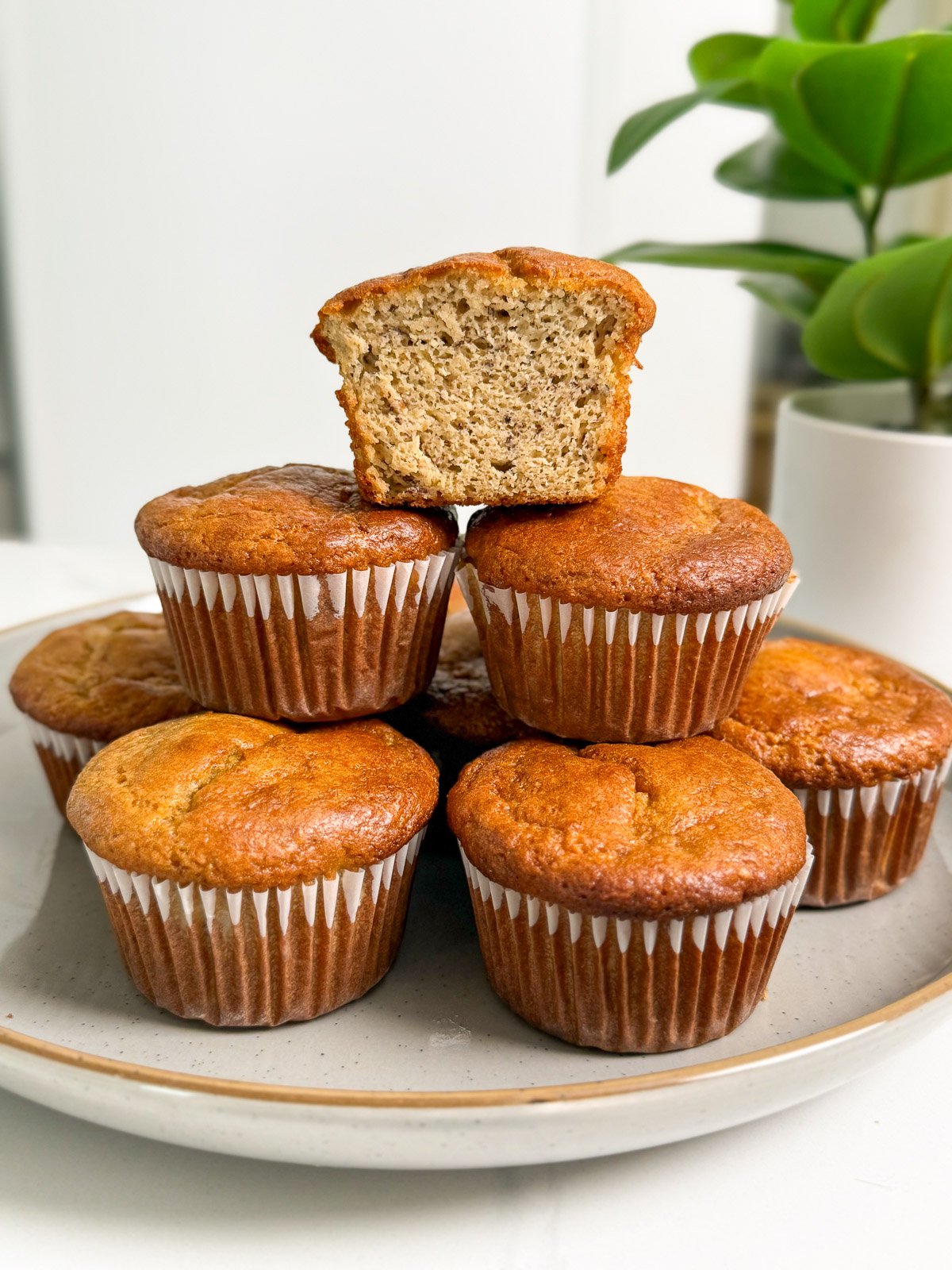A plate of six Protein Tahini Banana Muffins in white paper liners, with one muffin cut in half and placed on top to reveal the moist inside. A green potted plant sits in the background.