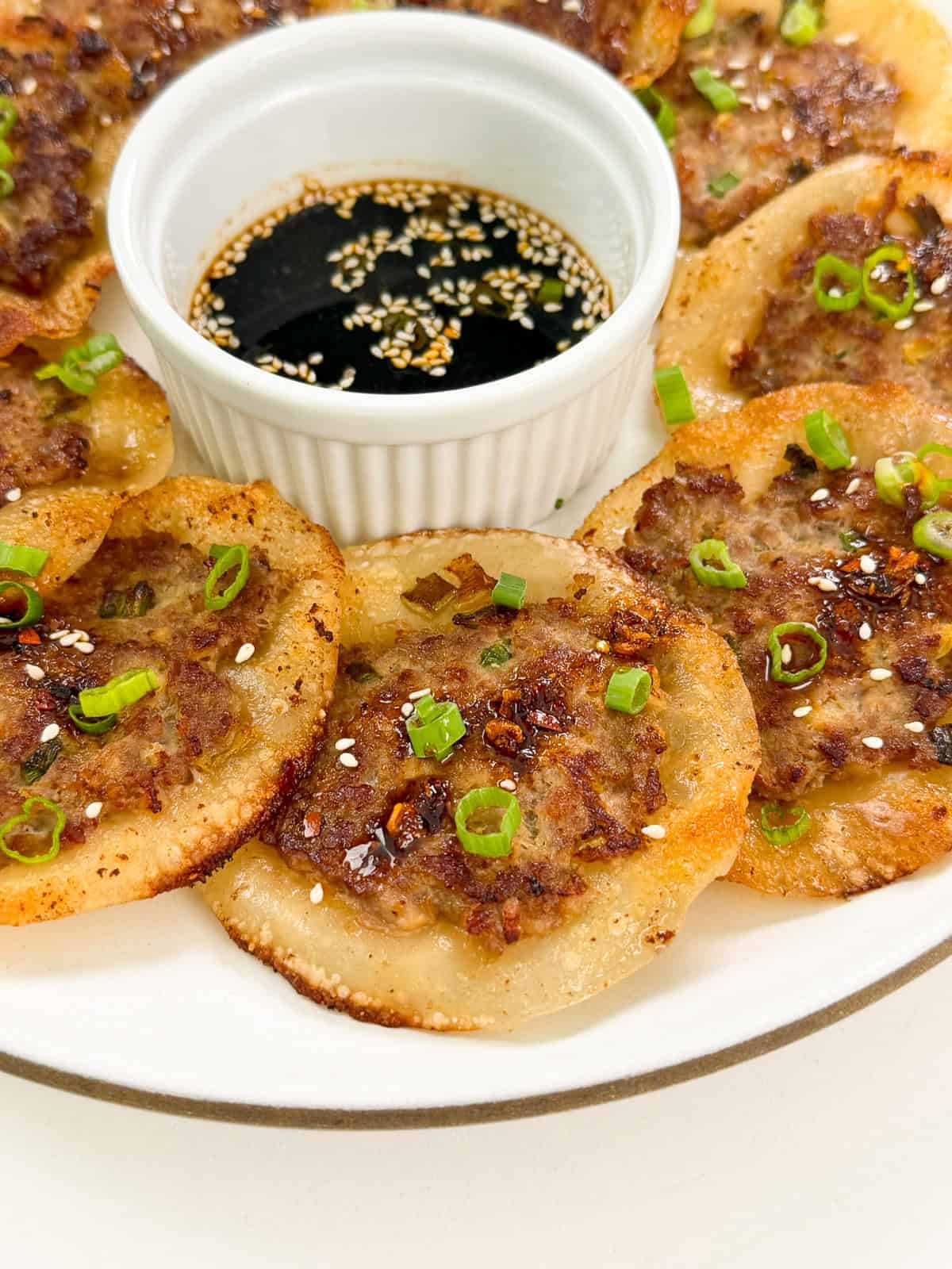 A plate of Smashed Lemongrass Pork Dumplings, pan-fried to perfection and garnished with chopped green onions and sesame seeds, arranged in a circle around a small dish of soy sauce with sesame seeds.