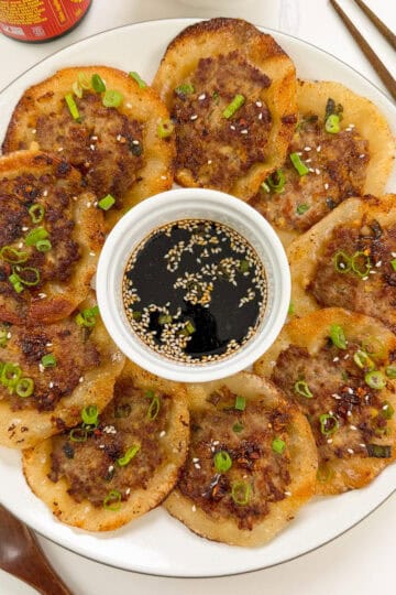 A plate of crispy Korean meat pancakes and smashed lemongrass pork dumplings, garnished with green onions and sesame seeds, arranged around a small bowl of soy dipping sauce. Chopsticks and a wooden spoon sit nearby.