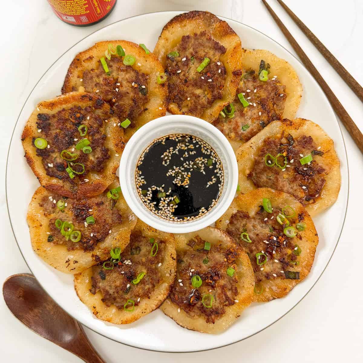 A plate of crispy Korean meat pancakes and smashed lemongrass pork dumplings, garnished with green onions and sesame seeds, arranged around a small bowl of soy dipping sauce. Chopsticks and a wooden spoon sit nearby.